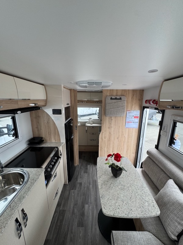 Standing at the bed looking towards the back of the caravan. On the right is the dining area, on the left the kitchen featuring a round stainless steel sink, cooktop and grill and the fridge/freezer. On the roof is the airconditioning unit and behind the dining area is the door to outside. At the far back is an internal doorway leading to the ensuite.