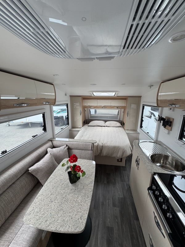 Standing at the door inside, looking towards the front of the caravan. On the immediate left is the dining seat (L shaped) and table, to the immediate right is the kitchen. In the back of the photo is a double bed, island configuration (can walk around both sides - N-S orientation).