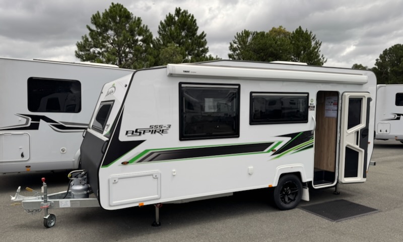 A white sandwich construction caravan with black and green markings down the side. There's a large sware window 1/3 in from the front, and a rectangular window behind that. Then the door just behind the axle. An awning is attached to the side above the windows and is in its stowed position in this photo. On the drawbar are two 4kg gas bottles.